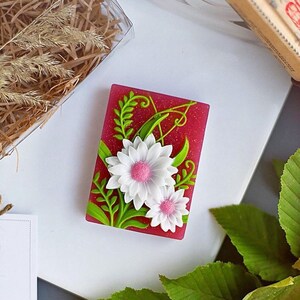 Puede incluir: Una pastilla de jab&oacute;n rosa con un dise&ntilde;o de flores blancas y rosas y hojas verdes. El jab&oacute;n tiene un acabado brillante.