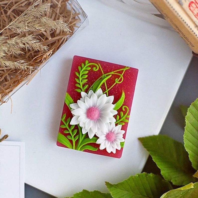 Puede incluir: Una pastilla de jab&oacute;n rosa con un dise&ntilde;o de flores blancas y rosas y hojas verdes. El jab&oacute;n tiene un acabado brillante.