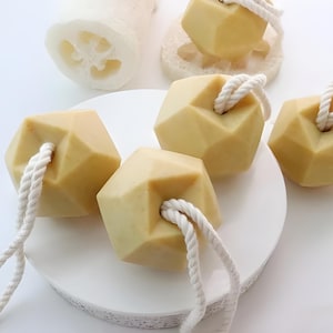 May include: Four beige geometric bath soap bars with rope handles. The soap bars are arranged on a white surface.