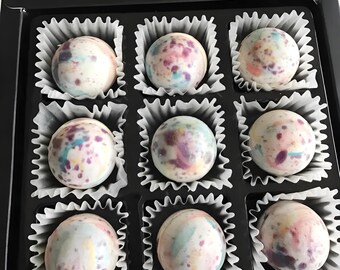 May include: A box of nine assorted chocolate truffles with colorful swirls and speckles. The truffles are arranged in a white paper-lined box with a black lid.