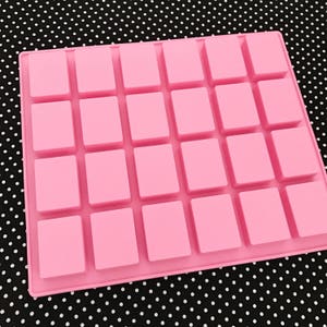K&ouml;nnte beinhalten: Eine rosa Silikonform mit einem Raster aus 28 rechteckigen Hohlr&auml;umen. Die Form ist f&uuml;r die Herstellung kleiner, quadratischer Gegenst&auml;nde konzipiert. Der Hintergrund ist ein schwarz-wei&szlig;es Polka-Dot-Muster.