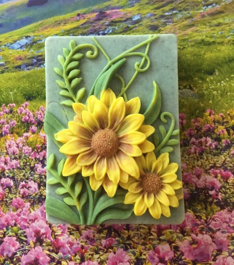 Puede incluir: Una pastilla de jab&oacute;n verde con un dise&ntilde;o en relieve de dos girasoles amarillos y hojas verdes.