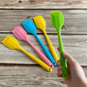 Könnte beinhalten: Eine Sammlung bunter Silikonspatel in Gelb, Rosa, Blau und Grün. Die Spatel haben einen langen, dünnen Griff und einen flachen, abgewinkelten Kopf. Ein Spatel wird von einer Hand gehalten. Die Spatel liegen auf einer Holzoberfläche.