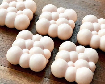 May include: A collection of light pink cloud-shaped items, each made up of rounded, bubble-like forms, are displayed on a wooden surface. The objects have a smooth, matte finish and are uniformly coloured. The wooden surface has a warm, brown tone.