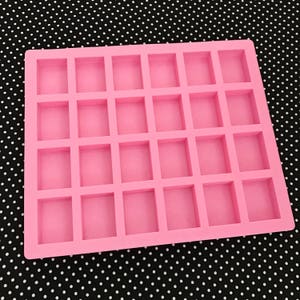 K&ouml;nnte beinhalten: Eine rosa Silikonform mit 30 rechteckigen Hohlr&auml;umen. Die Form dient zur Herstellung kleiner, quadratischer Gegenst&auml;nde wie Seifen, Kerzen oder Pralinen. Der Hintergrund ist ein schwarz-wei&szlig;es Polka-Dot-Muster.