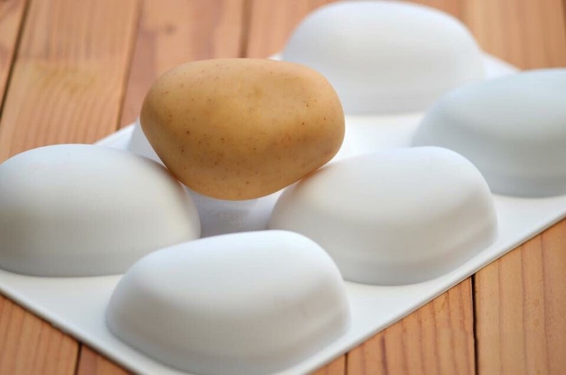 Puede incluir: Un molde de silicona blanco con seis cavidades con forma de piedras lisas y redondeadas. Una sola patata marr&oacute;n se encuentra en el centro del molde.