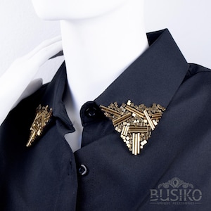 Può includere: Una camicia nera con colletto ornata da due spille geometriche dorate. Le spille presentano un mix di forme rettangolari e cilindriche, creando un design moderno e artistico. La camicia è abbottonata e presentata su un manichino.