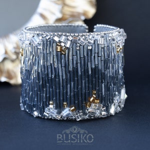 May include: A wide silver and gray beaded bracelet with gold accents. The bracelet is made of small, cylindrical beads and has a textured, geometric design.