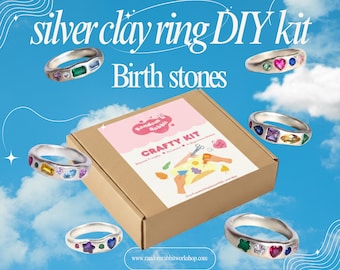 May include: A silver clay ring DIY kit featuring a brown cardboard box with a label that reads Random Rabbit Crafty Kit. Six silver rings with various colorful, multi-shaped gemstones are arranged around the box against a blue sky background.