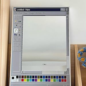 Könnte beinhalten: Ein rechteckiger Spiegel, der so gestaltet ist, dass er wie die Windows Paint-Programmoberfläche aussieht. Der graue Rahmen enthält eine Titelleiste, Menüoptionen und eine Farbpalette. Der Text "untitled - Paint" ist sichtbar. Der Spiegel steht auf einem Holzregal.