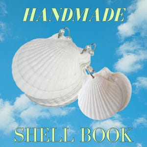 Puede incluir: Pendientes hechos a mano con forma de libro de conchas, con conchas blancas y aros plateados. Los pendientes se muestran sobre un fondo de cielo azul con nubes blancas. El texto "HANDMADE" y "SHELL BOOK" es visible en la imagen.