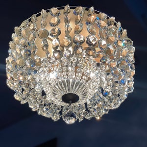 Peut inclure: Un lustre en cristal avec une base ronde argentée et de nombreux prismes en cristal transparent qui en pendent. Le lustre est allumé et diffuse une lueur chaude.