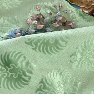 May include: A close-up of a light green fabric with a repeating pattern of stylized floral designs. The fabric has a textured, almost velvety feel. A decorative metal branch with clear glass flowers is placed on top of the fabric.