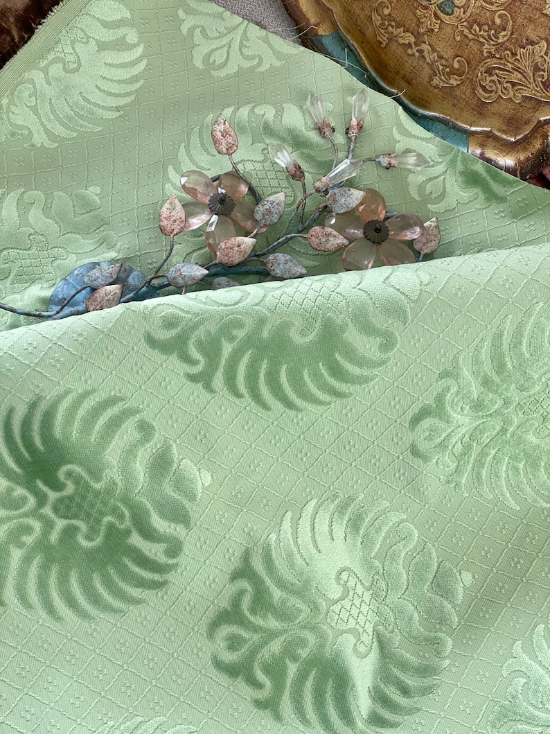 May include: A close-up of a light green fabric with a repeating pattern of stylized floral designs. The fabric has a textured, almost velvety feel. A decorative metal branch with clear glass flowers is placed on top of the fabric.