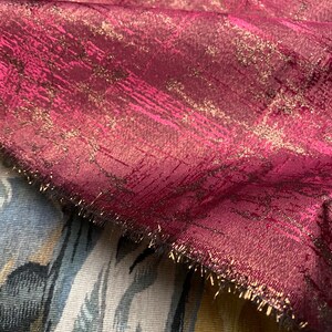 May include: Burgundy fabric with a gold metallic sheen and a textured pattern. The fabric has a fringed edge.
