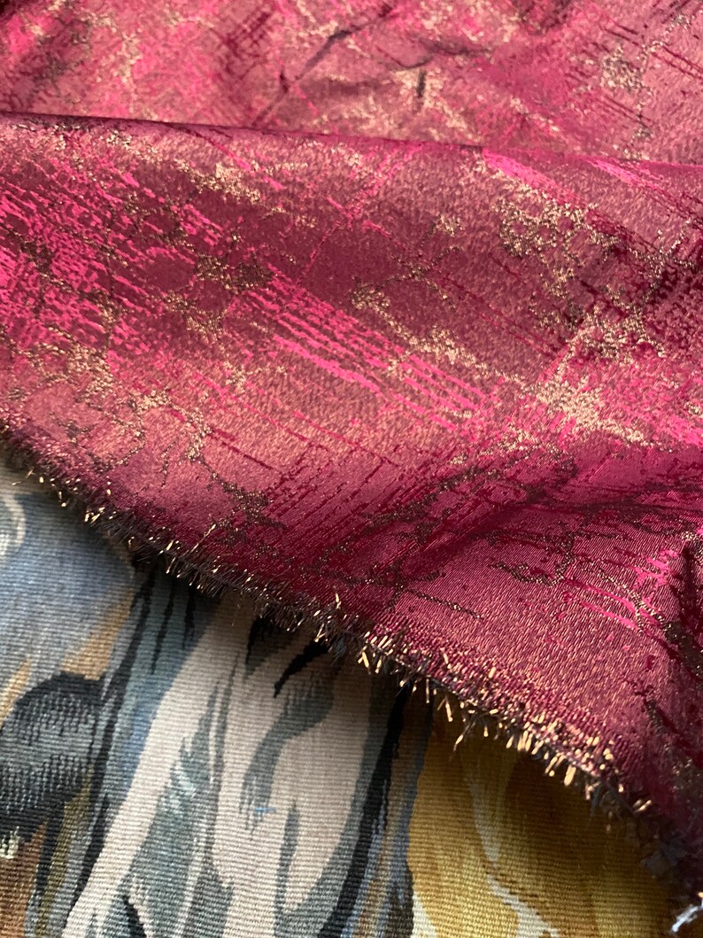 May include: Burgundy fabric with a gold metallic sheen and a textured pattern. The fabric has a fringed edge.