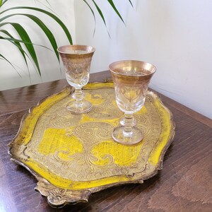 May include: A vintage serving tray with a bright yellow center and ornate gold trim. Two clear stemmed glasses with gold rims sit on the tray. The tray has a decorative, scrolled design.