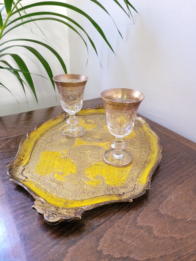 May include: A vintage serving tray with a bright yellow center and ornate gold trim. Two clear stemmed glasses with gold rims sit on the tray. The tray has a decorative, scrolled design.