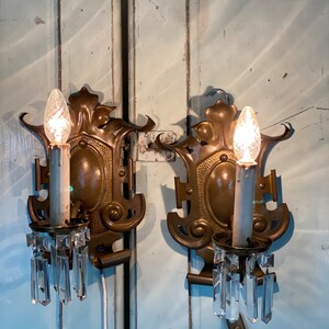 May include: A pair of antique brass wall sconces with crystal prisms. Each sconce features a decorative design with a central oval and ornate details. The sconces are mounted on a light blue wooden surface.