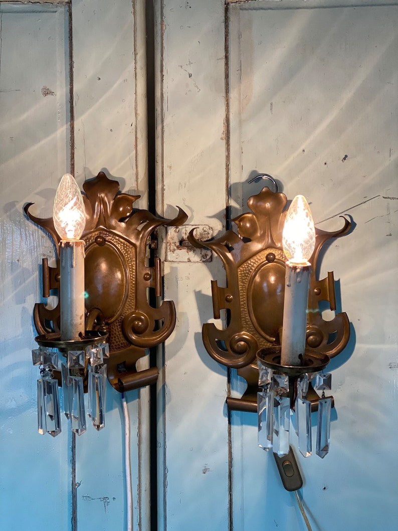 May include: A pair of antique brass wall sconces with crystal prisms. Each sconce features a decorative design with a central oval and ornate details. The sconces are mounted on a light blue wooden surface.