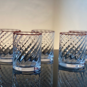 May include: Five clear glass tumblers with a red rim and a textured, swirling design. The glasses are arranged in a row on a black surface.