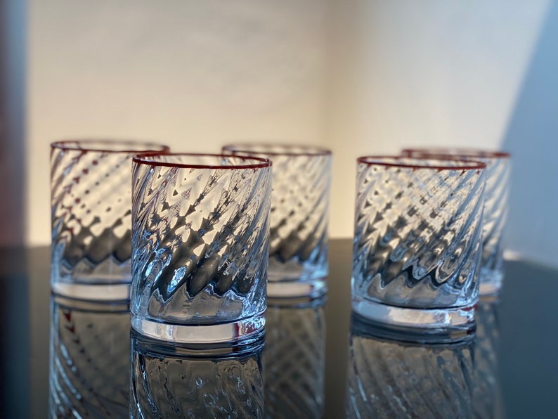 May include: Five clear glass tumblers with a red rim and a textured, swirling design. The glasses are arranged in a row on a black surface.