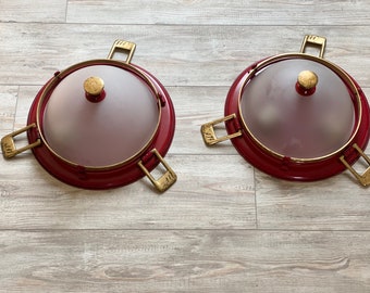 May include: Two red metal serving dishes with glass lids and gold handles. The dishes are on a light wood surface.
