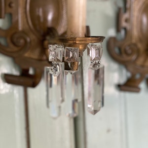 May include: A pair of antique brass wall sconces with crystal prisms hanging from the bottom. The sconces have a decorative design and are in good condition.