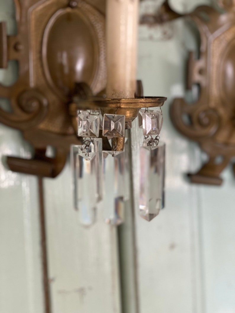 May include: A pair of antique brass wall sconces with crystal prisms hanging from the bottom. The sconces have a decorative design and are in good condition.