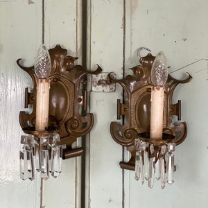 May include: A pair of antique brass wall sconces with crystal prisms. Each sconce features a candle-style bulb and intricate scrollwork.