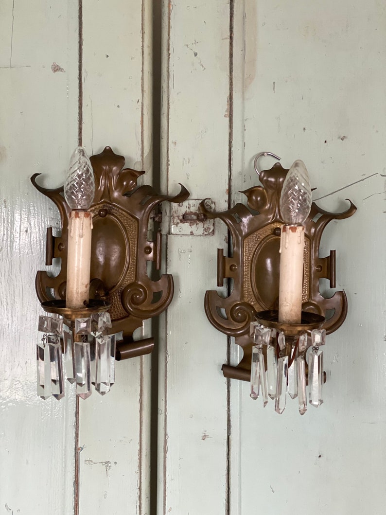 May include: A pair of antique brass wall sconces with crystal prisms. Each sconce features a candle-style bulb and intricate scrollwork.