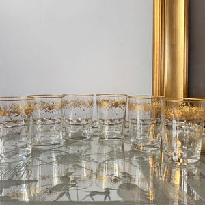 May include: Set of eight clear glass tumblers with gold trim and a gold floral design. The glasses are arranged in two rows of four on a reflective surface.