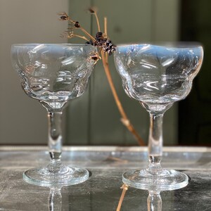 Pu&ograve; includere: Due calici trasparenti con bordo decorativo, affiancati. Tra di loro c'&egrave; un rametto di fiori e bacche secche. I calici sono su una superficie riflettente.