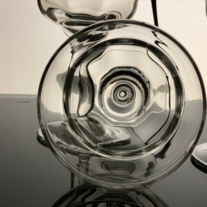 Pu&ograve; includere: Bicchieri di vetro trasparente con una coppa arrotondata e un lungo stelo. L'immagine mostra il lato inferiore del bicchiere, rivelando cerchi concentrici e un design dettagliato. I bicchieri sono disposti su una superficie riflettente.