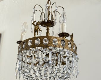 May include: A brass chandelier featuring a circular frame with embossed patterns, adorned with multiple clear glass crystals in various shapes. The fixture includes small brass cherub figures, white candle-style light covers, and a chain.