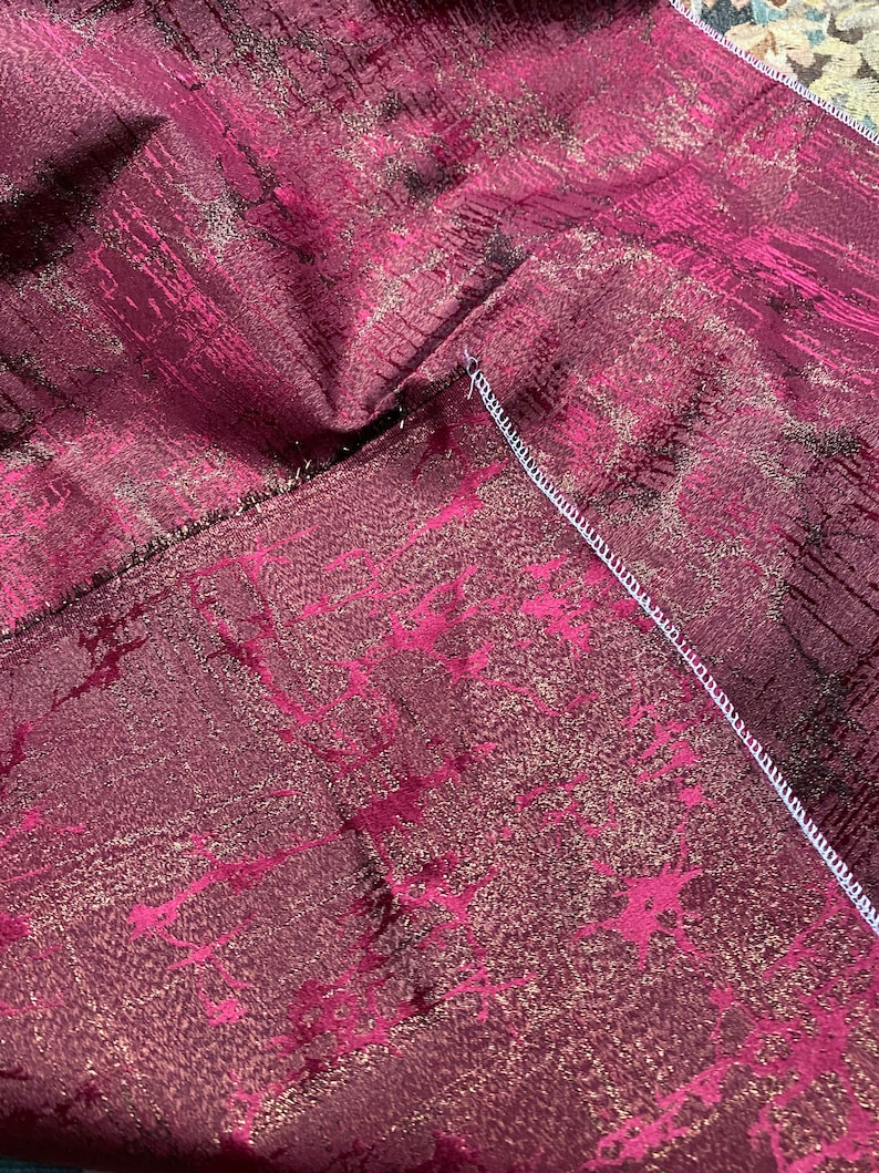 May include: Close-up of a burgundy fabric with a gold metallic pattern. The fabric has a subtle sheen and a textured surface.