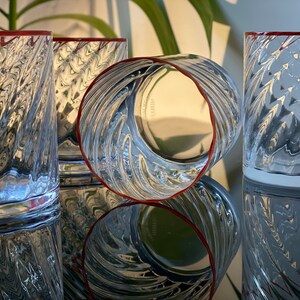 May include: Four clear glass tumblers with a textured, wavy design. The glasses have a red rim and are arranged in a row on a reflective surface.