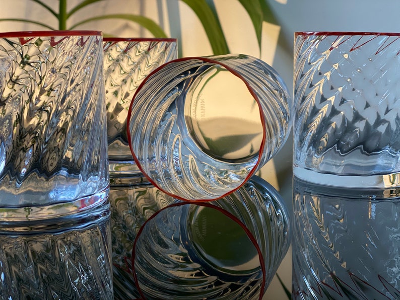 May include: Four clear glass tumblers with a textured, wavy design. The glasses have a red rim and are arranged in a row on a reflective surface.