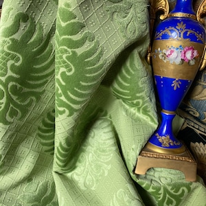 May include: A green velvet fabric with a damask pattern. The fabric is draped over a blue and gold decorative vase with floral details.