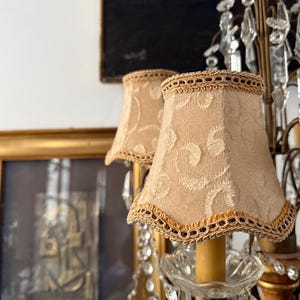 May include: A close-up of a vintage chandelier featuring two small, beige lampshades. The lampshades have a delicate floral pattern and are trimmed with gold and burgundy accents. The chandelier has crystal pendants and a gold frame.