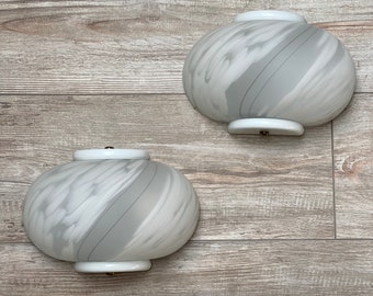 May include: Two white glass wall sconces with a swirl pattern. The sconces are mounted on a light wood background.