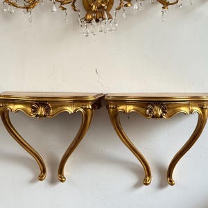 May include: Two ornate, gold-colored console tables with curved legs and a shell-shaped design on the front. The tables have a light-colored top and are mounted on a white wall. A gold chandelier with crystal accents hangs above.