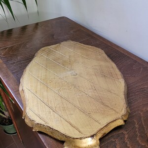 May include: A gold-colored, leaf-shaped tray with a textured surface and decorative edges. The tray has a distressed finish, and is resting on a wooden surface.