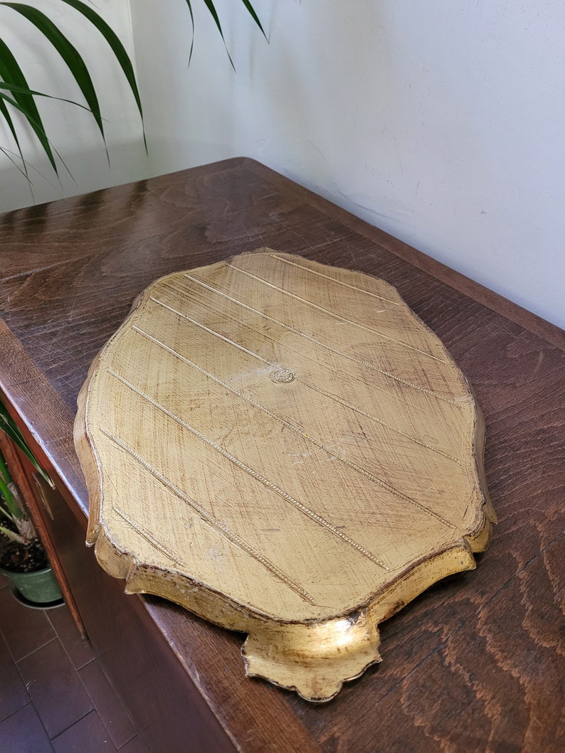 May include: A gold-colored, leaf-shaped tray with a textured surface and decorative edges. The tray has a distressed finish, and is resting on a wooden surface.