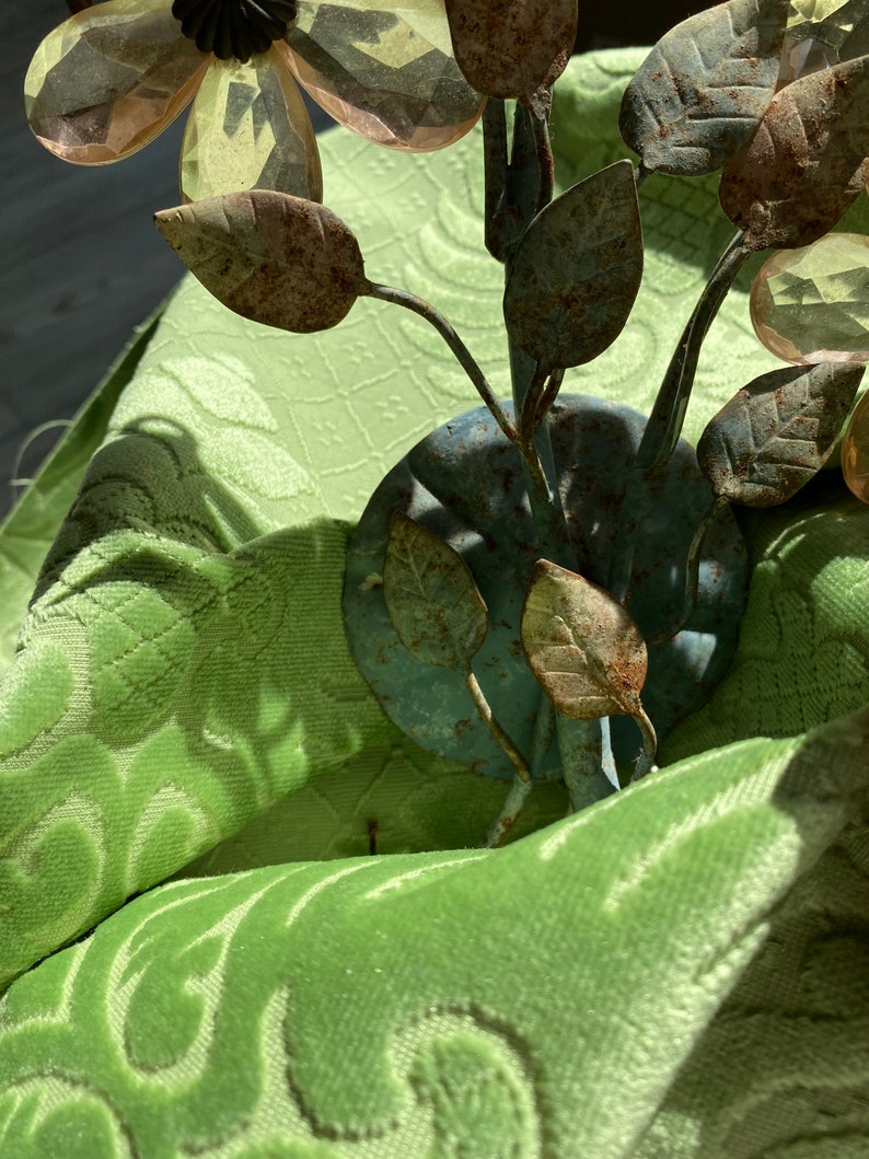 May include: A metal flower sculpture with rust-colored leaves and clear glass flowers. The sculpture is sitting on a green fabric surface.