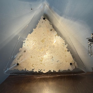May include: A triangular wall lamp featuring a clear glass frame with a textured, shattered white glass inlay. The fixture is secured by three small black circular fasteners at the corners, emitting a warm, diffused light through the centre.