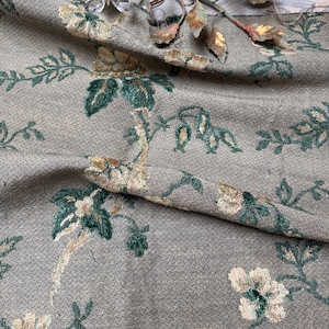 以下が含まれることがあります： ベージュ、グリーン、クリーム色の花柄が施された、質感のある生地のクローズアップ。生地はドレープされており、デザインの詳細が明らかになっています。背景には、クリスタルのアクセントが付いた装飾的な金属片が見えます。
