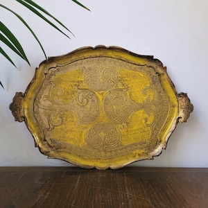 May include: A decorative, antique-style serving tray with a yellow and gold finish. The tray has an ornate, scalloped edge and features intricate embossed floral designs. Two handles are on either side. The tray is on a wooden surface.