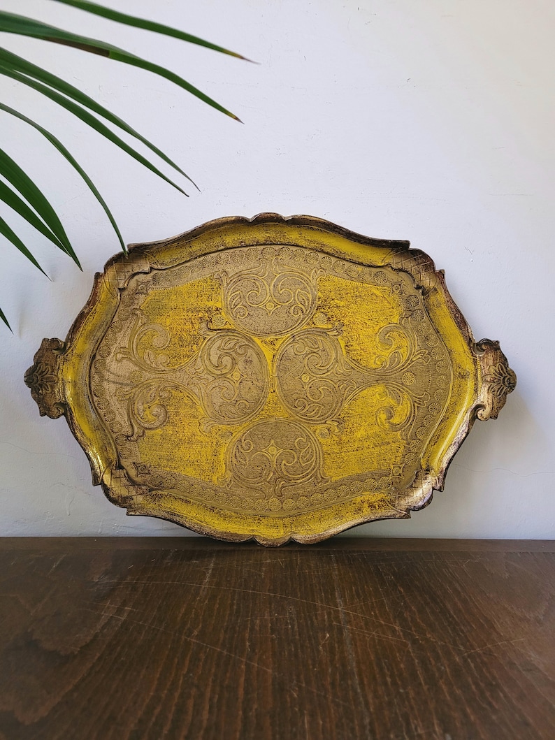 May include: A decorative, antique-style serving tray with a yellow and gold finish. The tray has an ornate, scalloped edge and features intricate embossed floral designs. Two handles are on either side. The tray is on a wooden surface.