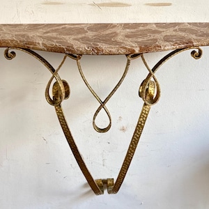 May include: A wall-mounted console shelf featuring a polished brown and white marble top with a smooth, curved edge. The support structure is crafted from hammered gold-toned metal, showcasing intricate scrollwork and a V-shaped frame.